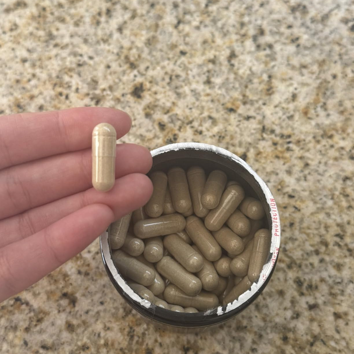 Hand holding a supplement capsule above a container of capsules on a granite countertop.
